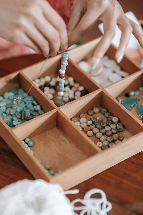 Crop unrecognizable female stringing beads on elastic cord while making bracelet at table with kit of assorted supply in room