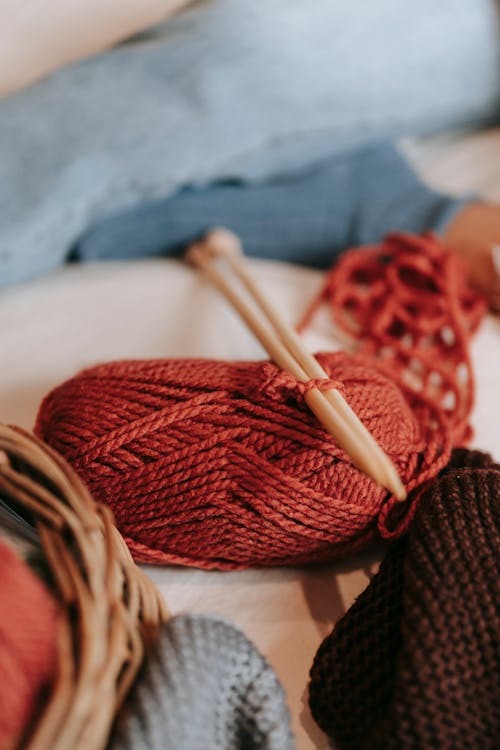 Skein of red yarn with wooden knitting needles placed on soft couch with crop faceless person in light room at home