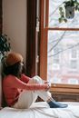 Side view of dreamy young African American female in warm clothes sitting on bed while looking away against window