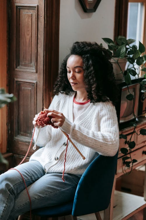 Attentive young African American craftswoman with curly hair and yarn knitting with needles in light house room