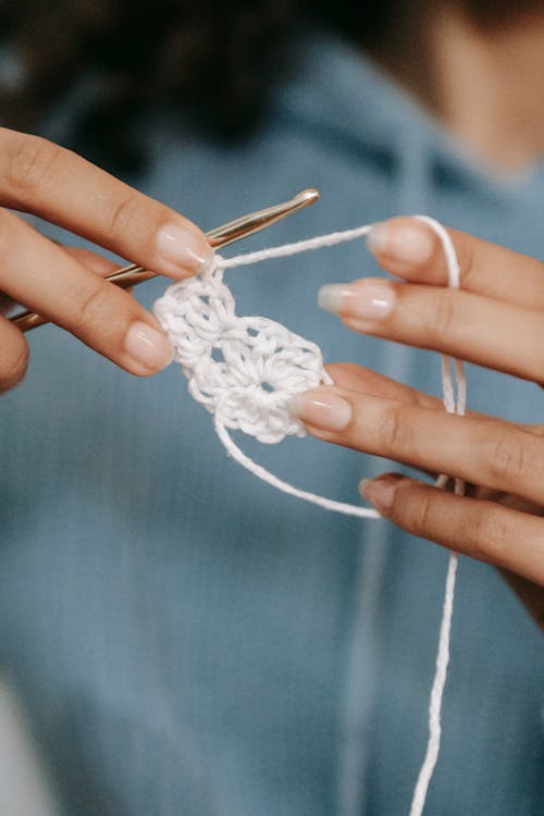 Crop anonymous female artisan with hook and white yarn crocheting flower while having spare time on blurred background