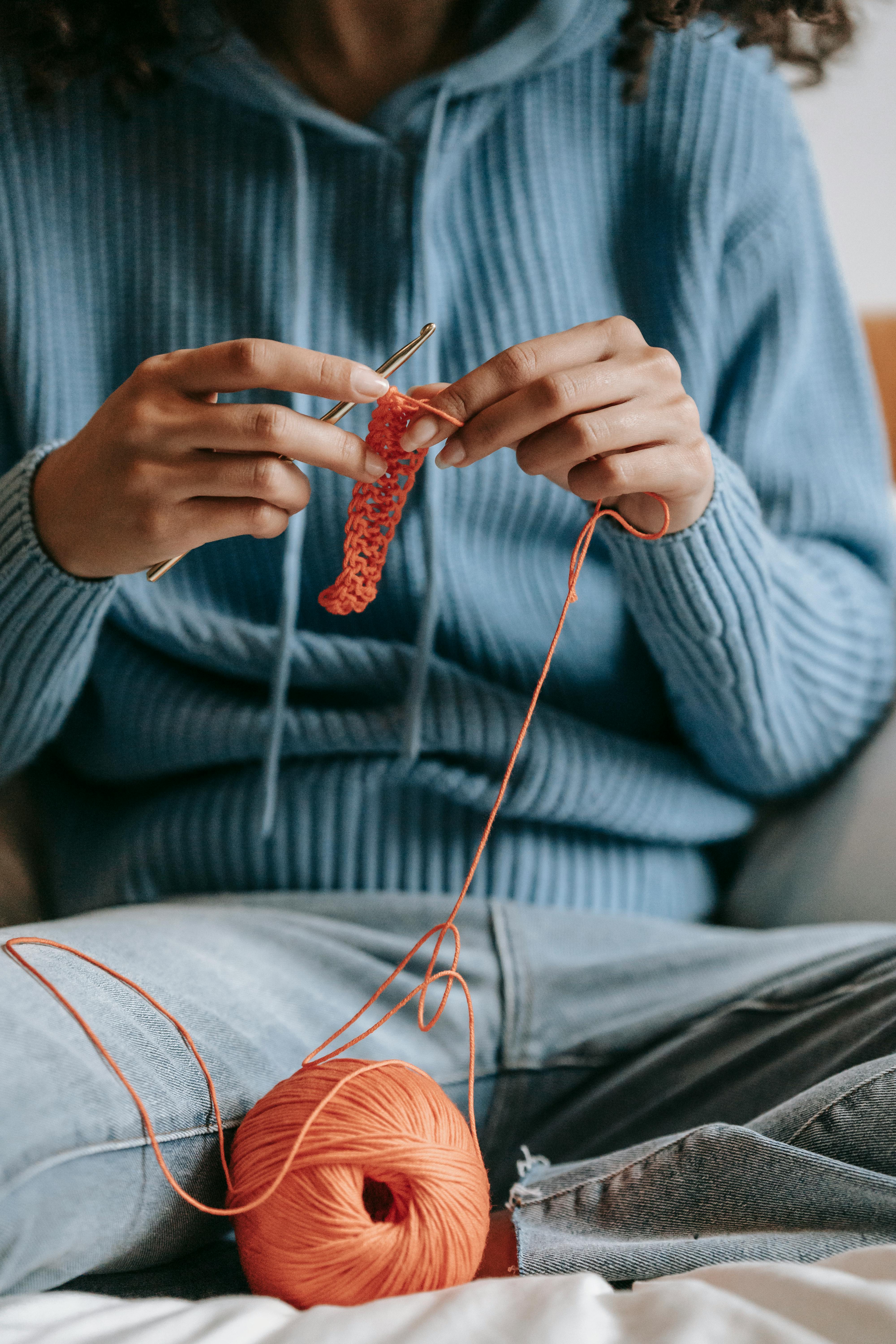 Crop unrecognizable female artisan with hook and yarn ball crocheting with crossed legs on bed in house