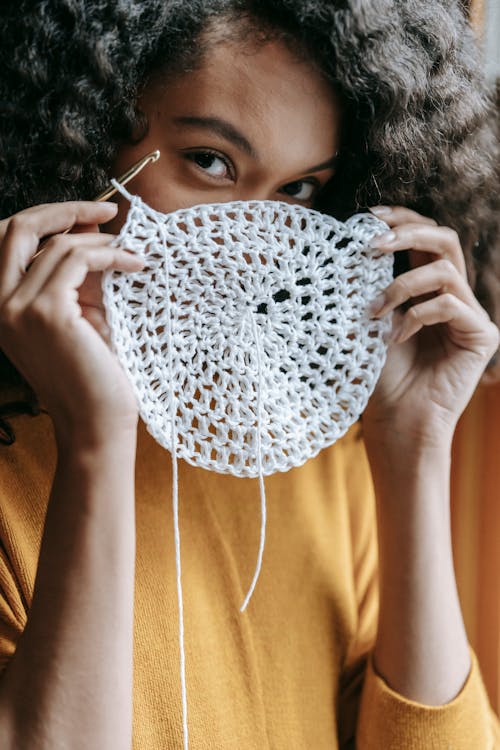 Free African American craftswoman looking at camera while showing crocheted fabric and hook in house room Stock Photo