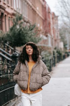 Young African American female with curly hair and hands in pockets looking at camera in town