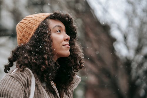 Free Side view of dreamy young black female in knitted hat looking up in town on snowy day Stock Photo