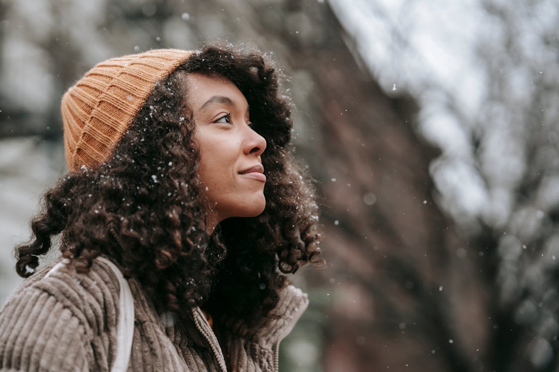 Side view of dreamy young black female in knitted hat looking up in town on snowy day