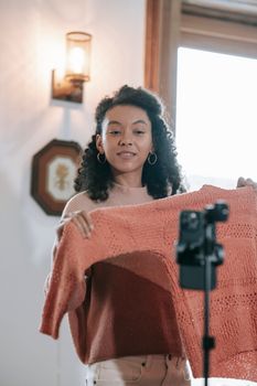 African American female vlogger with handmade knitted sweater recording video on cellphone in house room