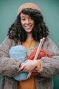 Young positive ethnic female with dark curly hair wearing light orange knitted hat and light brown corduroy jacket with skein of yarn and knitting needles