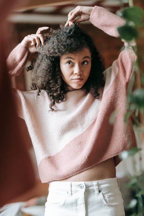 Positive young African American lady with dark curly hair wearing light pink sweater and white jeans doing hairstyle at mirror