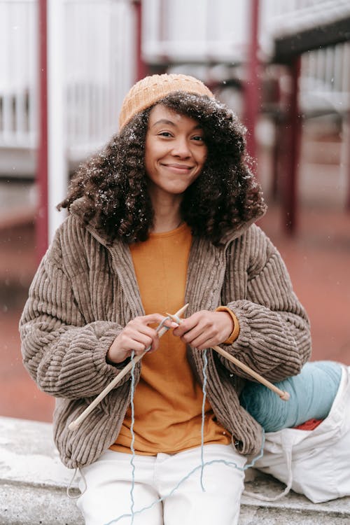 Positive African American woman with dark lush hair wearing orange pullover and beige jacket and white jeans hat doing needlework