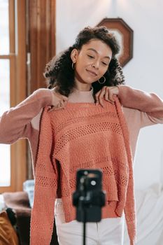 Smiling young African American woman with curly hair trying on orange sweater and taking selfie photo on phone fixed on tripod