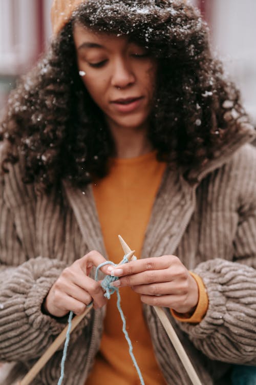 Concentrated African American female with snowflakes on curly dark hair wearing yellow sweater and brown jacket knitting with blue woolen yarn and needles outdoors
