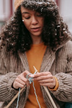 Concentrated African American female with snowflakes on curly dark hair wearing yellow sweater and brown jacket knitting with blue woolen yarn and needles outdoors