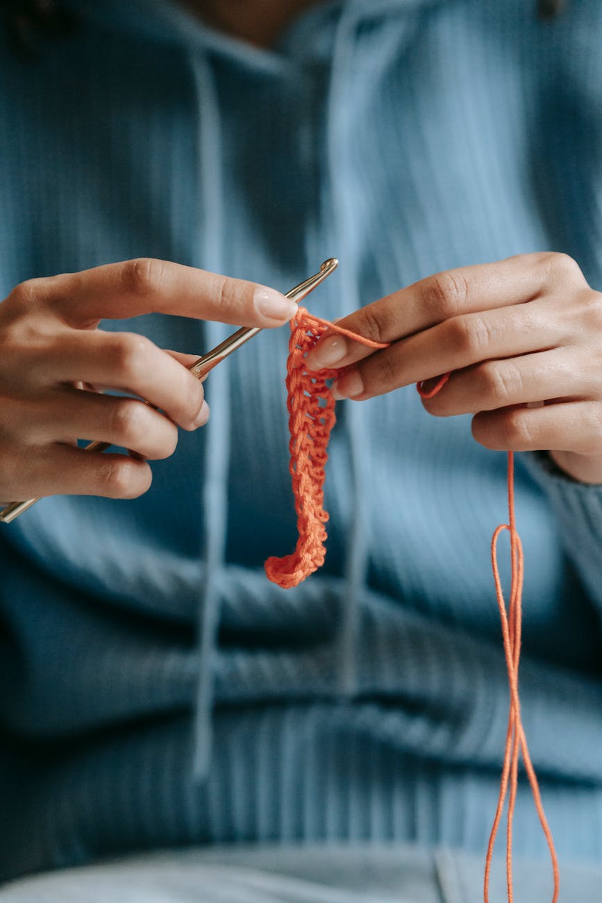 Crop faceless female in warm blue pullover knitting with orange threads and crochet.