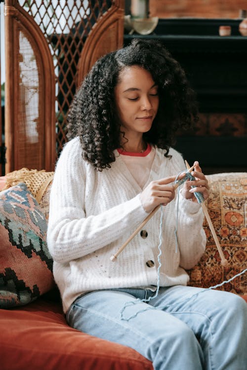 Concentrated black lady knitting with blue yarn using wooden needles while sitting on soft couch