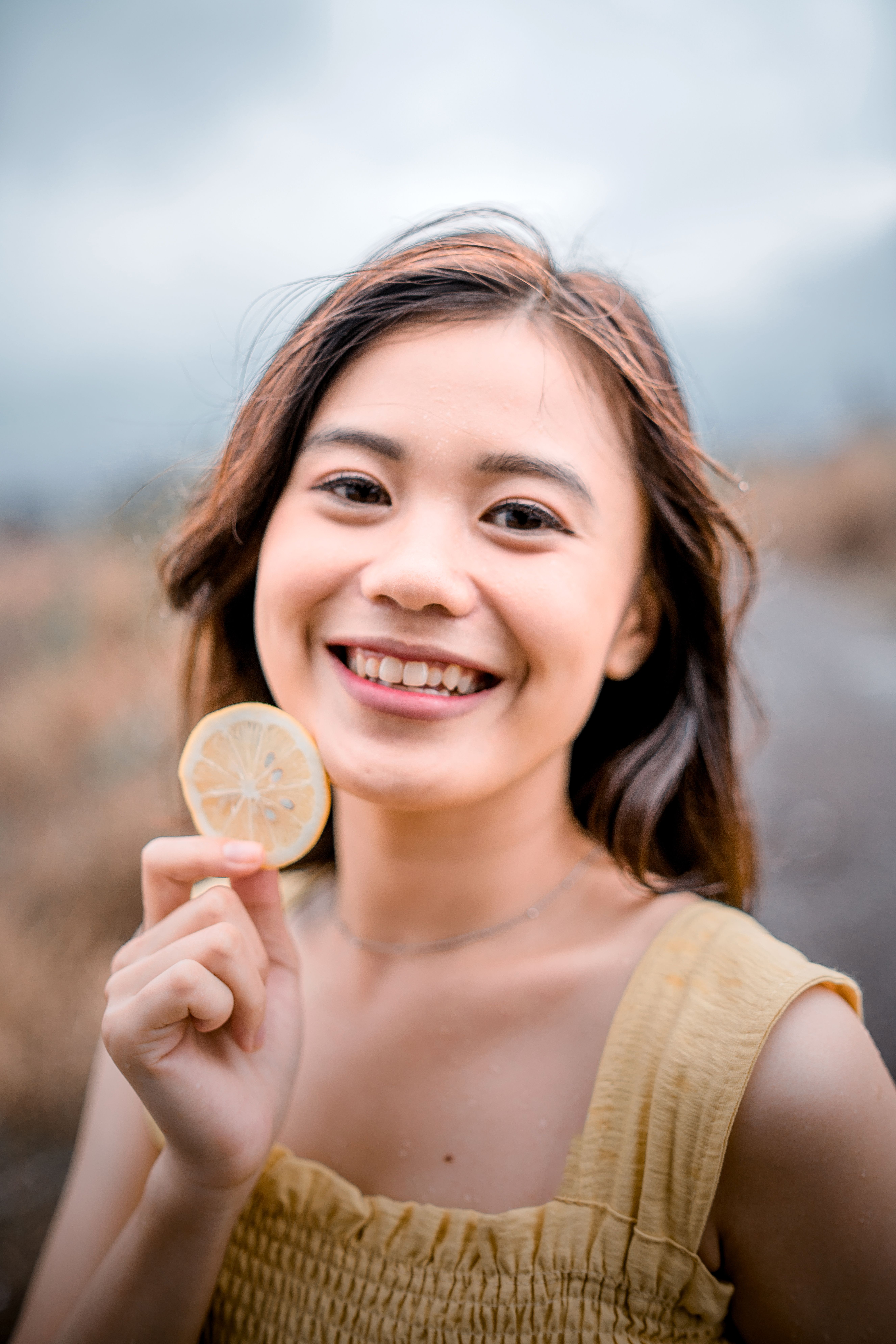 A smiling young woman holding a lemon slice poses in a natural outdoor setting.