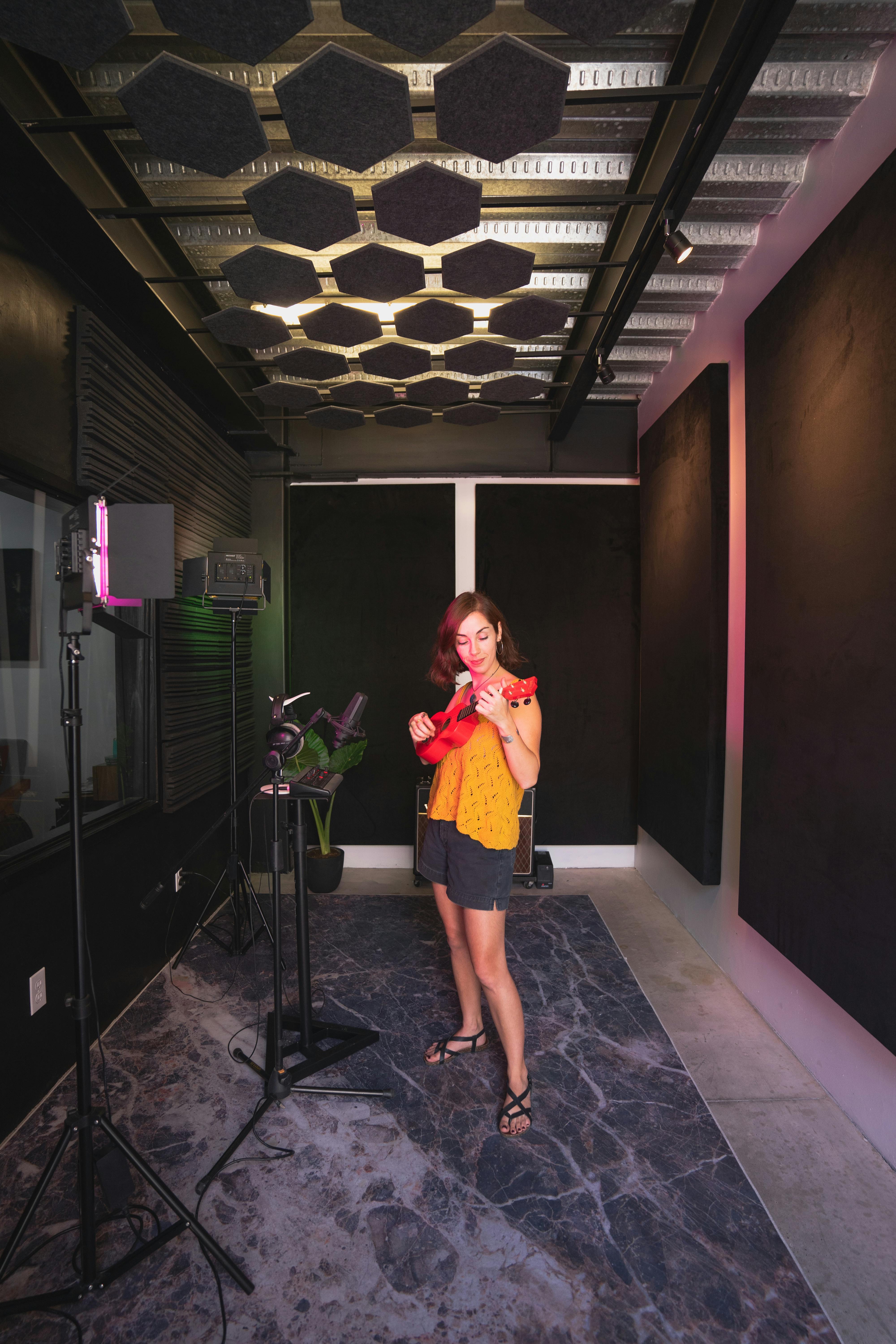 A woman strums a ukulele in a modern recording studio filled with sound equipment.