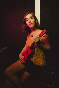 A young woman sits indoors playing a red ukulele, creating a vibrant musical scene.