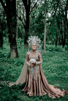 A bride stands elegantly in a traditional wedding dress holding a bouquet in a lush forest setting.