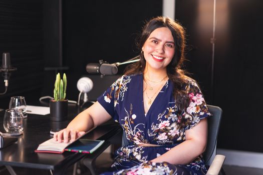 Woman in a floral dress sitting in a podcast studio, smiling with a microphone in view.