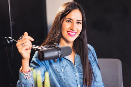 A woman in a denim shirt, smiling while podcasting in a studio with a professional microphone.
