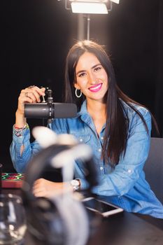 A cheerful woman wearing a denim shirt sits in a recording studio with a microphone.