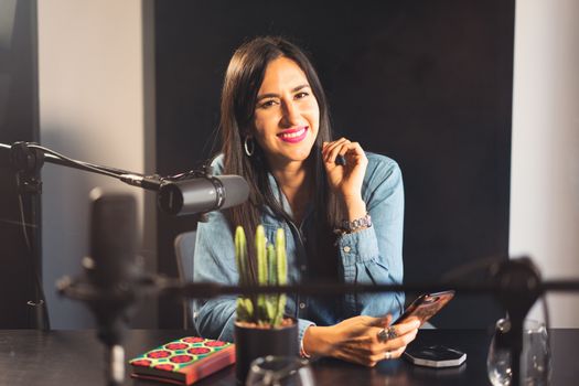 Smiling woman in denim recording a podcast with microphone, phone, and cactus on table.