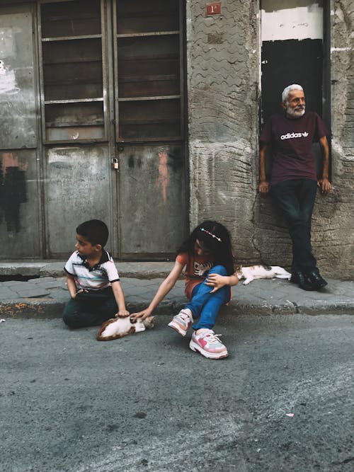 Full body of ethnic kids sitting on asphalt walkway near senior man standing at shabby building on street in town