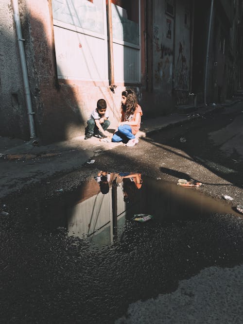 Full body of ethnic local children sitting on narrow sidewalk near road with puddle in sunny poor district of town
