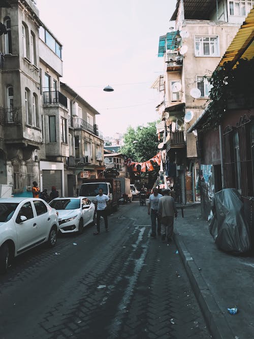 People walking on roadway with automobiles between old residential houses located on street in district of town on summer day