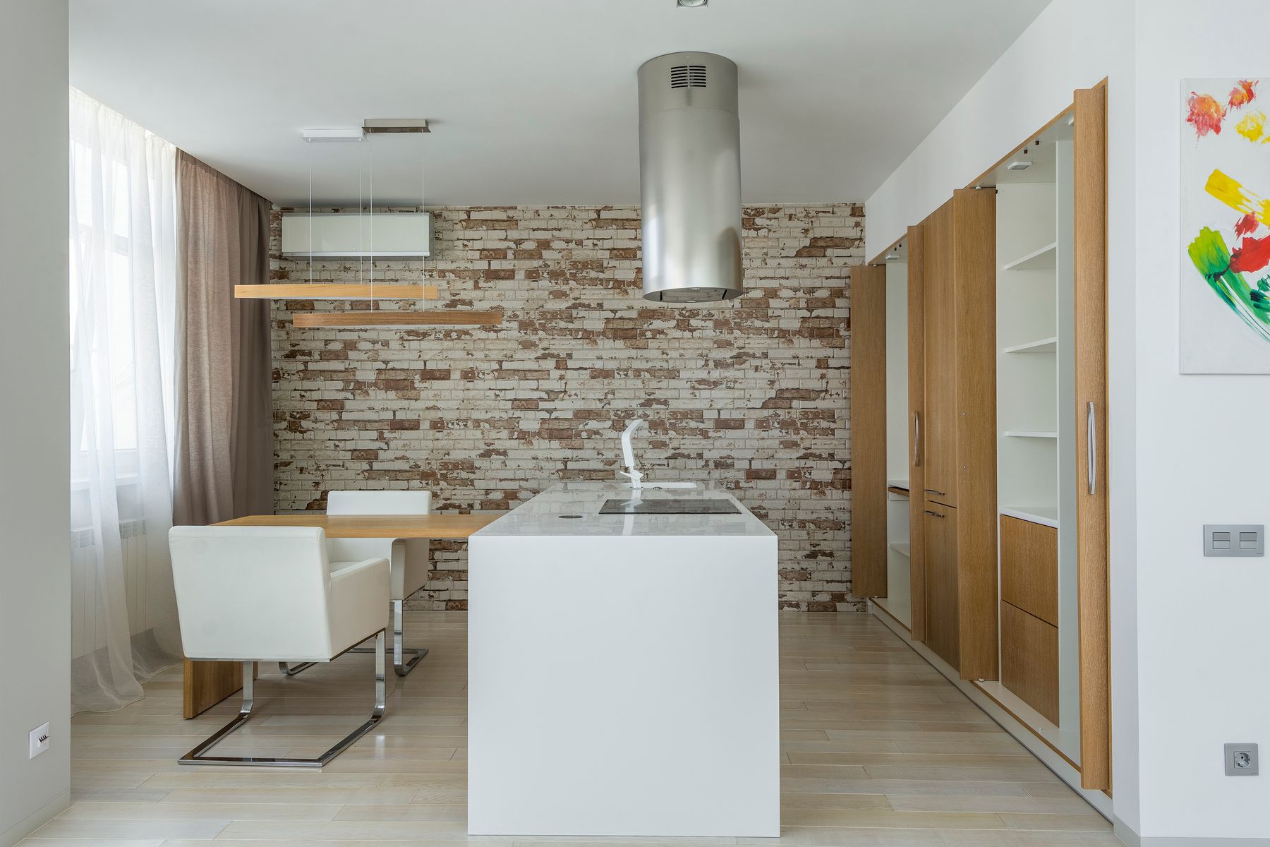 White kitchen island and dining chairs with a pale brick wall, wood cabinets and metal extractor