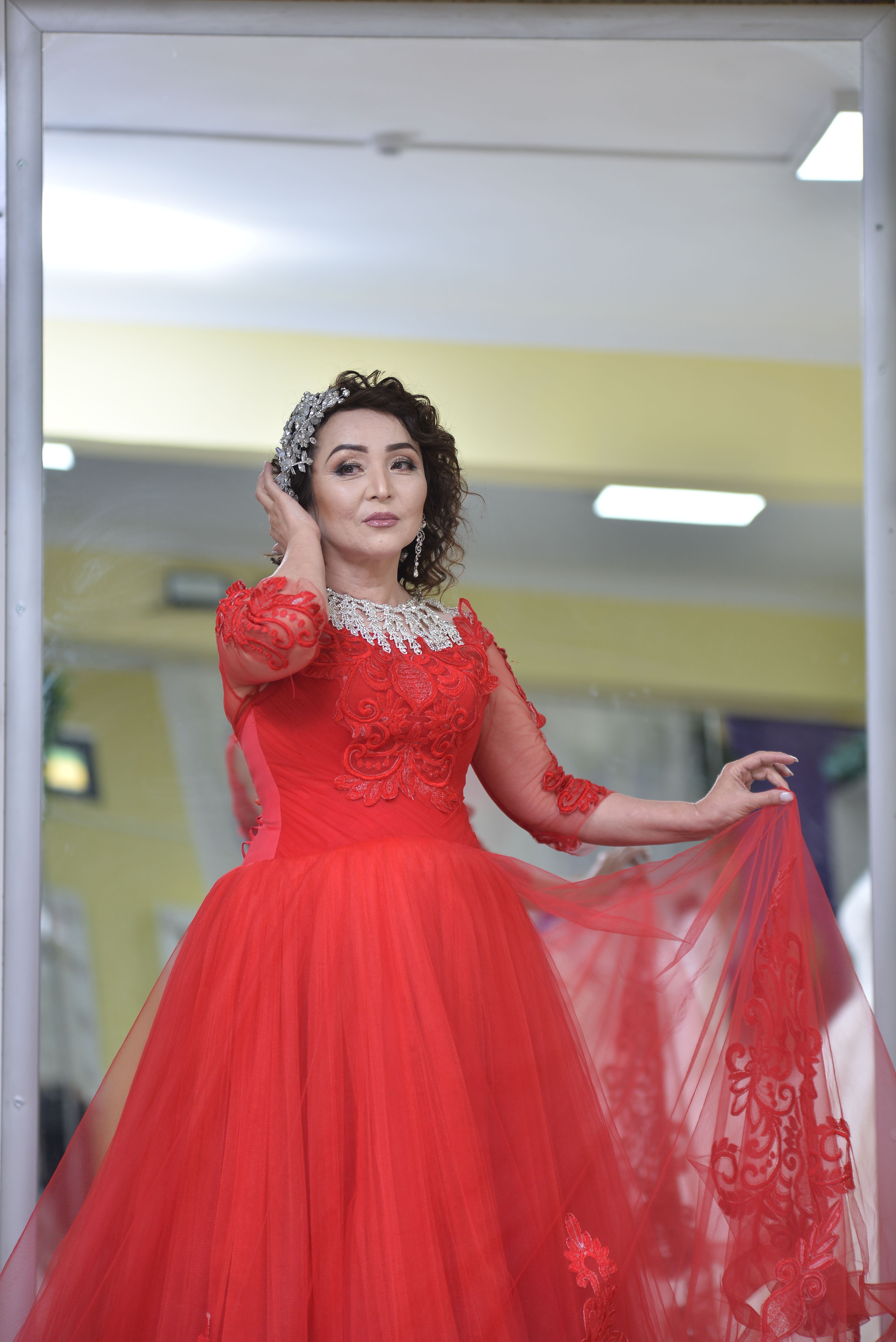 Woman in an elegant red gown posing indoors, showcasing sheer fabric with intricate lace details.