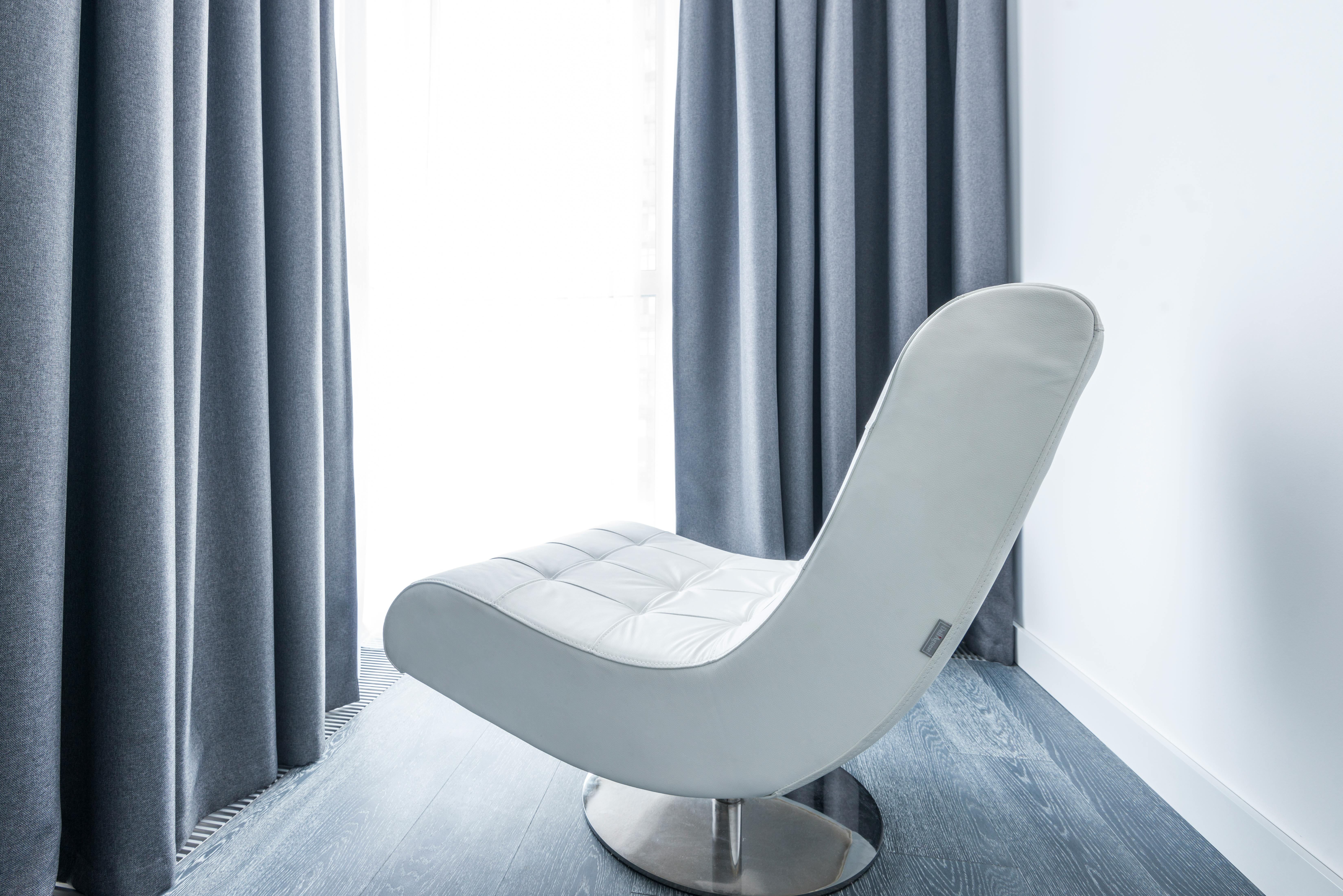 Free A modern white leather lounge chair in a minimalist room with grey drapes and natural lighting. Stock Photo