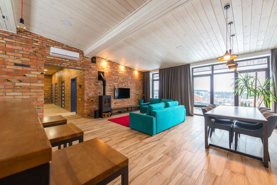 Photo by Max Vakhtbovych on Pexels Spacious modern living room featuring a brick wall, teal sofa, and large windows.