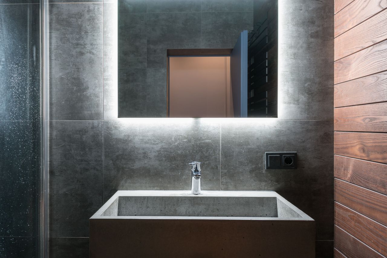 Concrete sink under a backlit mirror in a dark-tiled bathroom