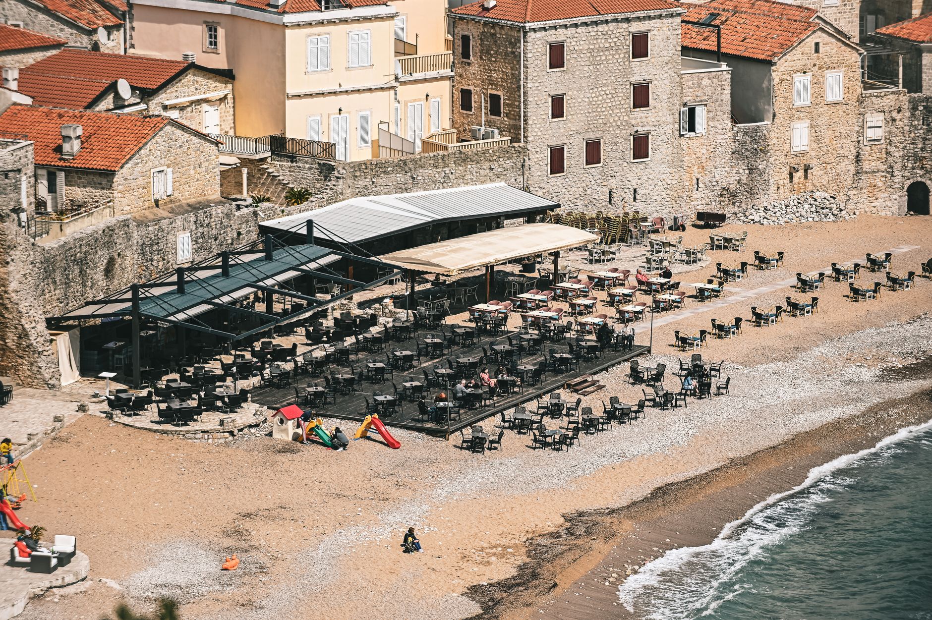 Aerial view of an empty beachfront café and historic stone buildings by the seashore.