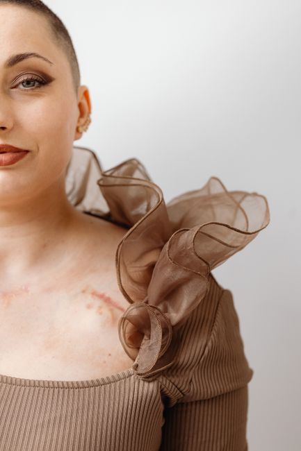 Close-up of a stylish woman with a prominent scar and elegant, unique fashion details in a soft, minimalistic setting.