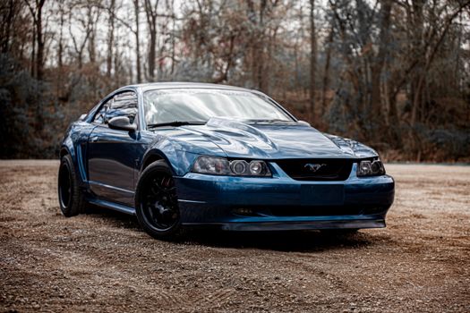A striking blue Ford Mustang parked on a gravel path with a forest backdrop.