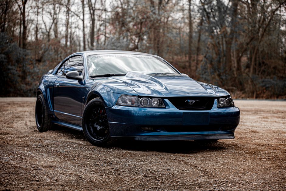 A striking blue Ford Mustang parked on a gravel path with a forest backdrop.