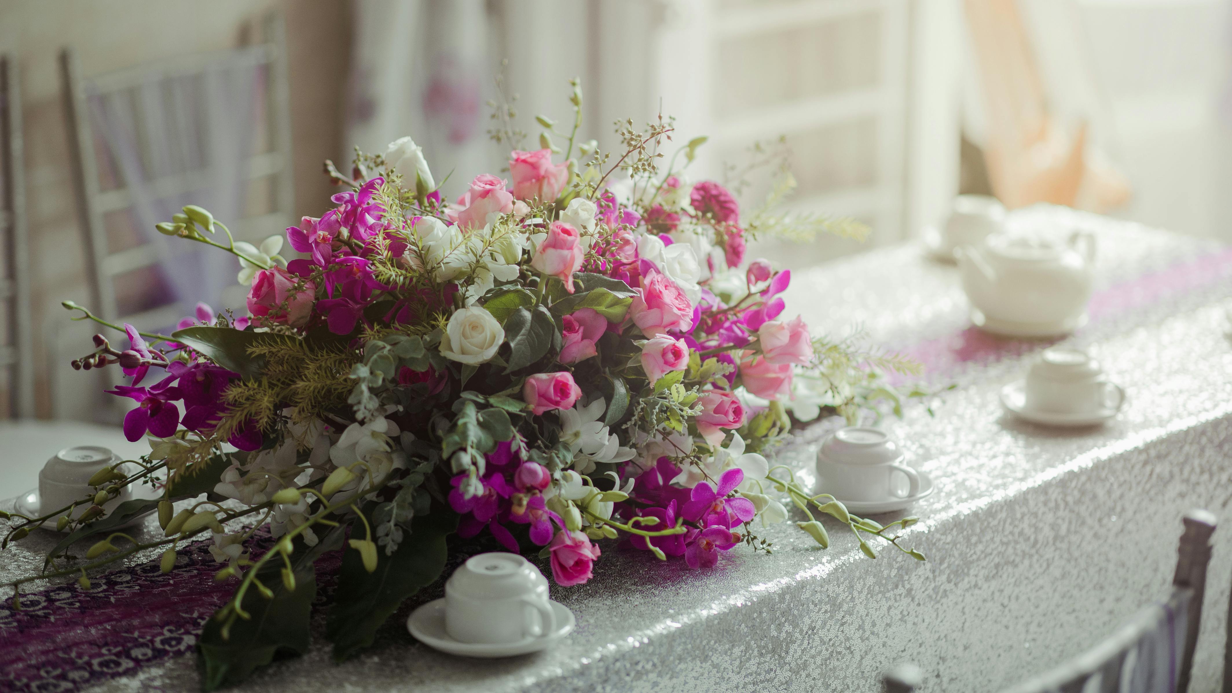 餐桌上摆放着粉白相间的美丽花卉，上面还有白色的茶杯