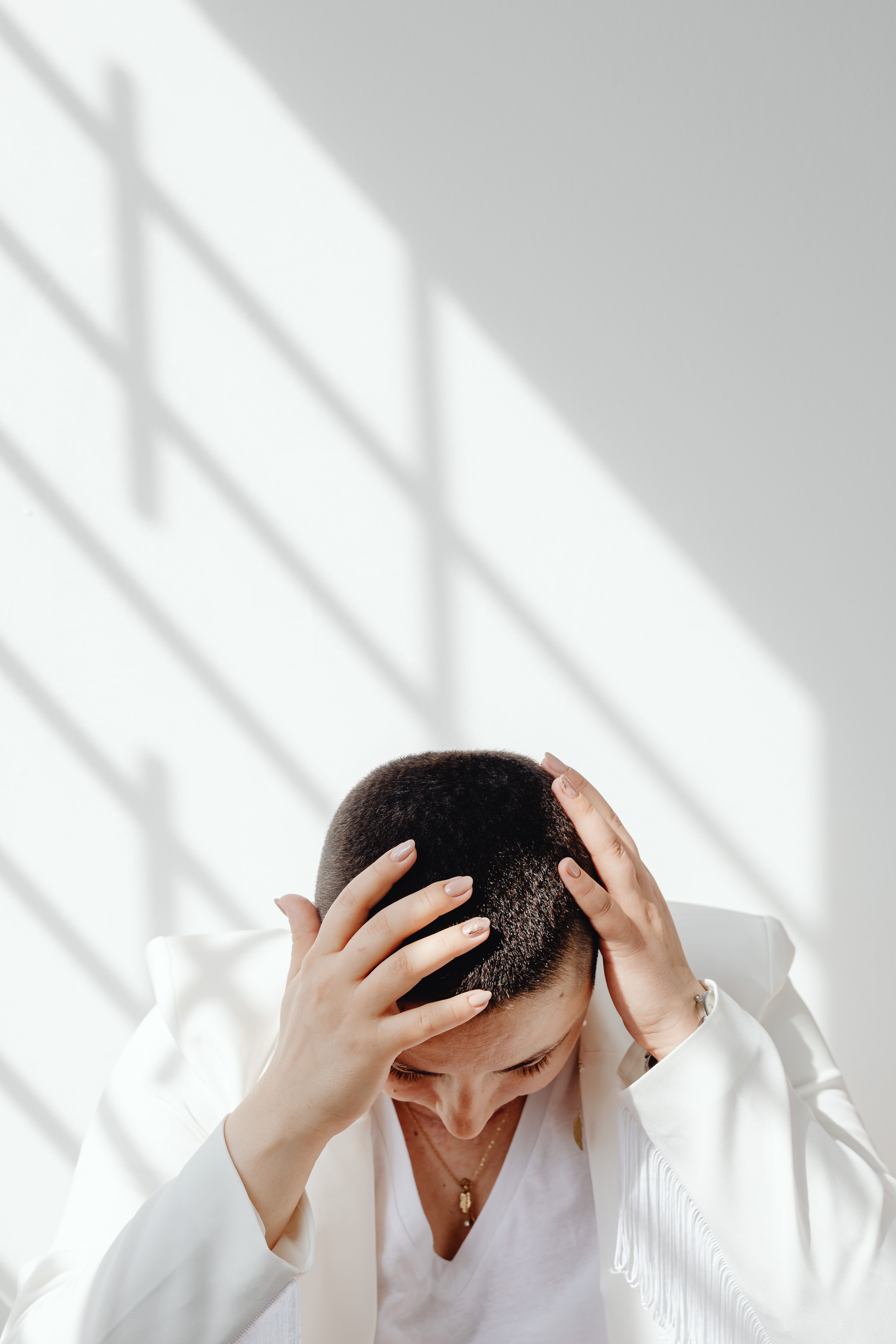 A woman appears stressed, holding her head while light streams in through modern window blinds.