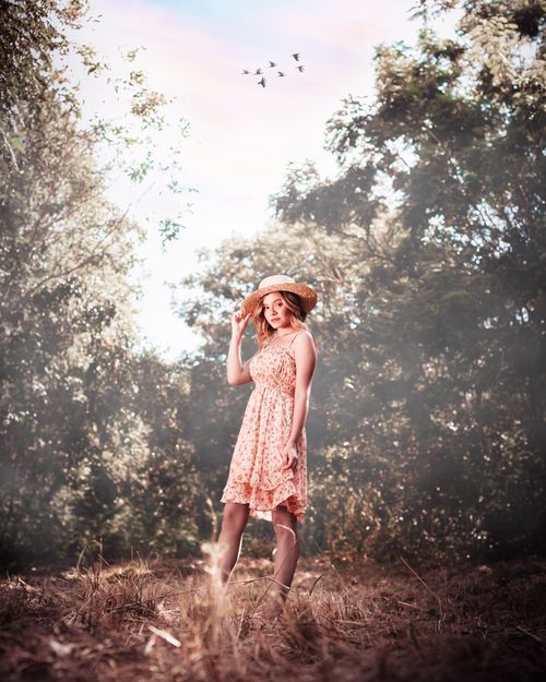 Free A woman in a floral dress poses with a straw hat in a sunlit forest. Stock Photo