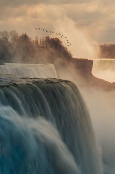 Golden sunset over Niagara Falls, capturing mist and a flock of birds in motion.