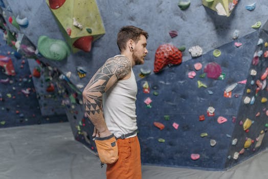 A tattooed man with a chalk bag taking a break in an indoor climbing gym.