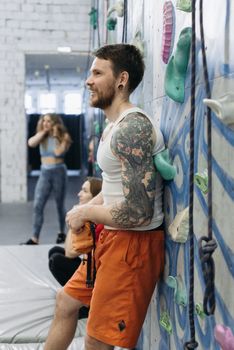Tattooed man in tank top rests against climbing wall indoors, showcasing athletic lifestyle.