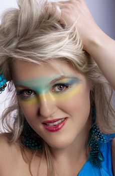 Woman with vibrant makeup, feather earrings, and stylish hair, posing confidently indoors.