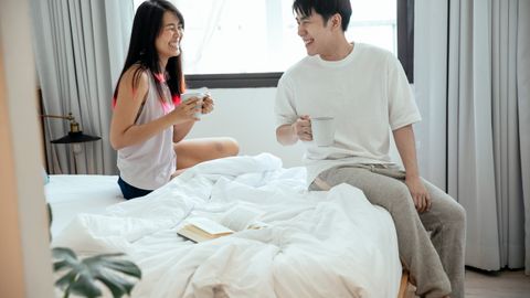 Laughing Asian couple looking at each other and enjoying hot coffee in morning while sitting on bed