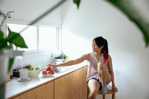 Free Positive ethnic young lady in pajama sitting near counter with utensil and apples in plate near window in kitchen in daytime Stock Photo