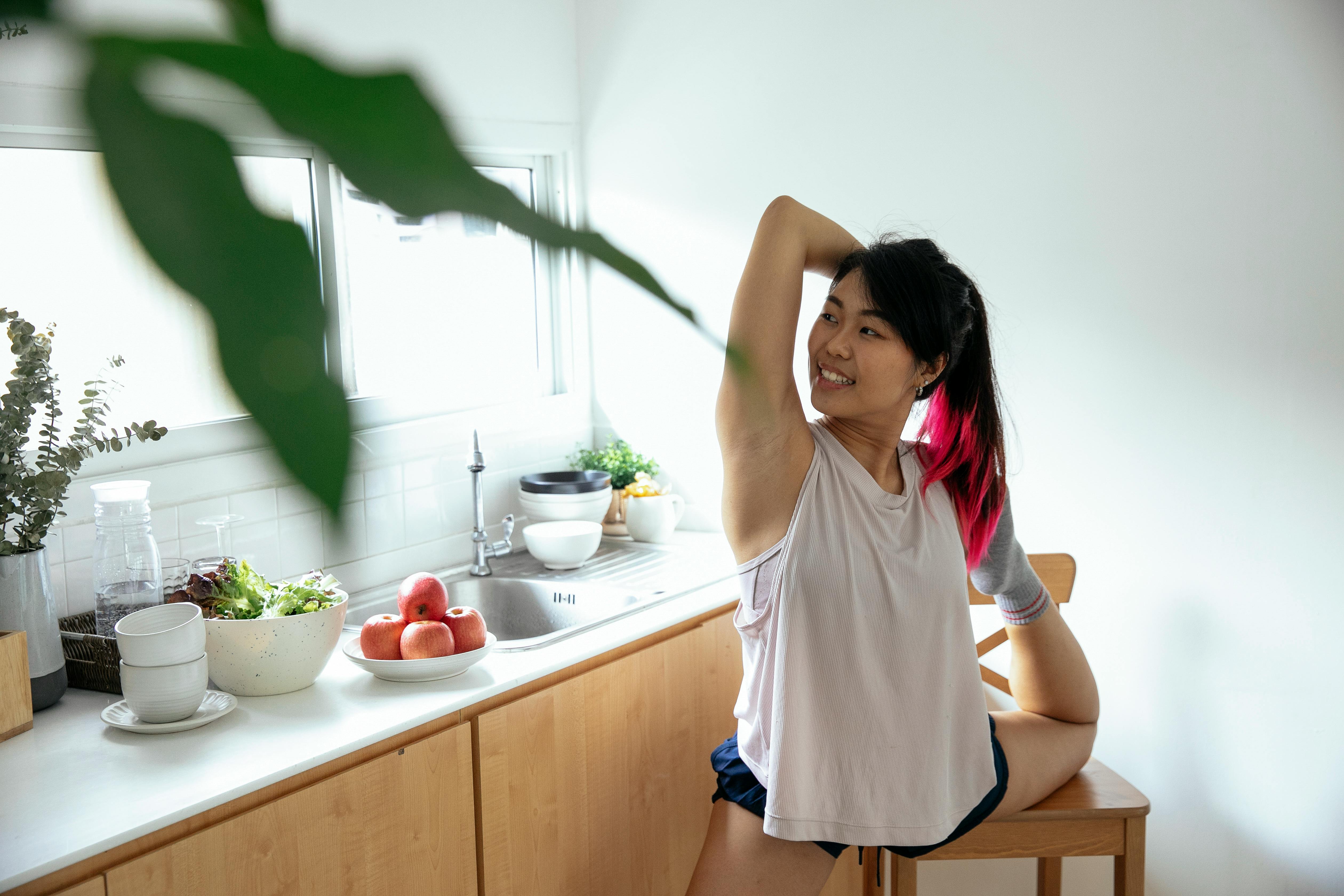 smiling asian woman doing standing bow asana in kitchen