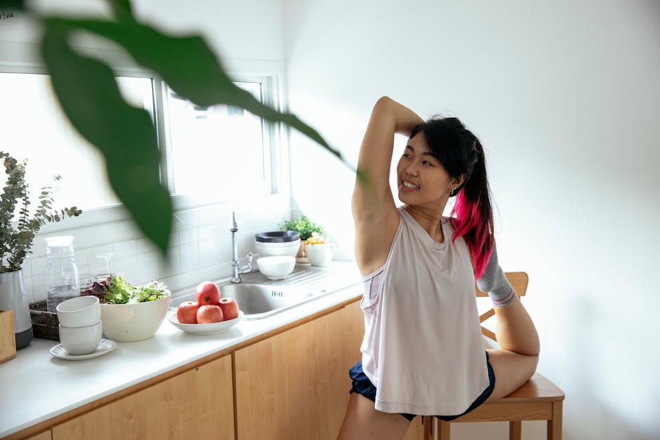 Cheerful Asian female in casual clothes stretching leg using chair while practicing Standing Bow yoga asana in kitchen